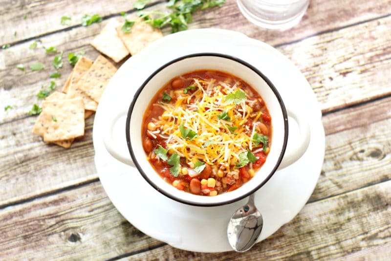 White bowl containing a southwestern style beef stew with tomatoes, beans and corn, topped with shredded cheese, crackers and cilantro on table. 