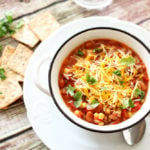 White bowl containing a southwest stew with tomatoes, beans and corn, topped with shredded cheese, crackers and cilantro on table.