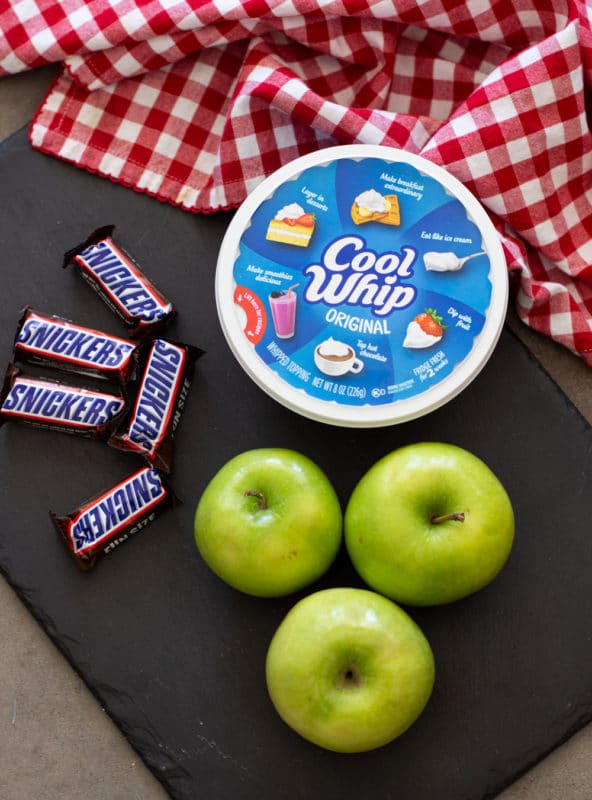 Cool whip, granny smith apples, and snickers bars on a black cutting board.