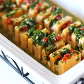 White glass dish containing orange and white marinated cheese topped with parsley, pimentos and garlic.