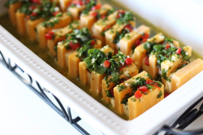 White glass dish containing orange and white marinated cheese topped with parsley, pimentos and garlic.