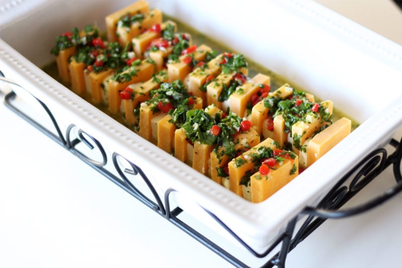 White glass dish on a black wire stand containing sharp cheddar cheese and cream cheese topped with parsley, pimentos, olive oil and garlic.