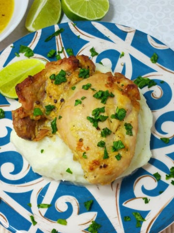 Blue and white plate containing Baked Chicken with Dijon Lime Sauce sitting on a bed of mashed potatoes topped with fresh parsley, lime wedges on side.