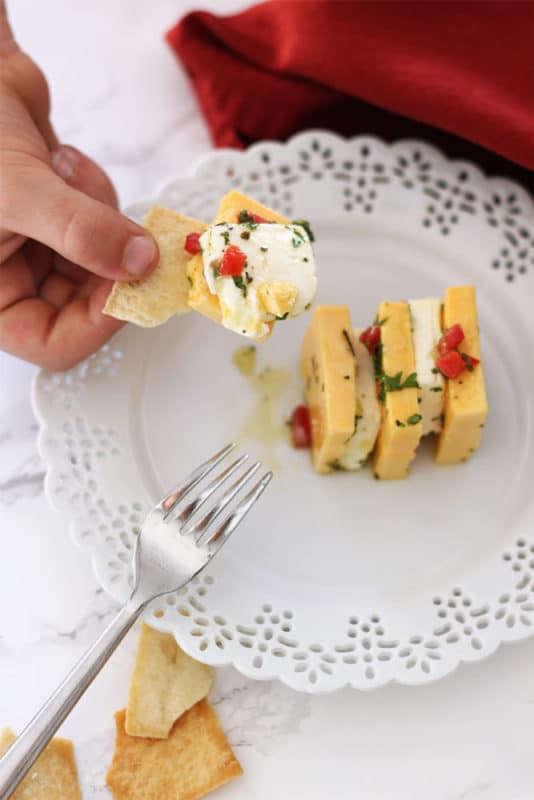 White lace plate with pita chips on the side, hand holding a pita chip covered with sharp cheddar and cream cheese, topped with pimentos, parsley, olive oil and garlic.