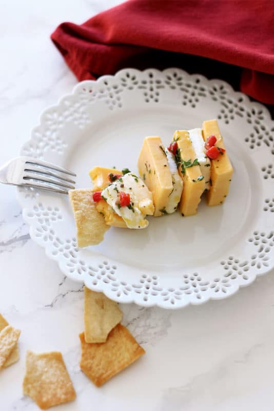 White lace plate containing cream cheese and cheddar cheese served with pita chips, red napkin and fork in background.