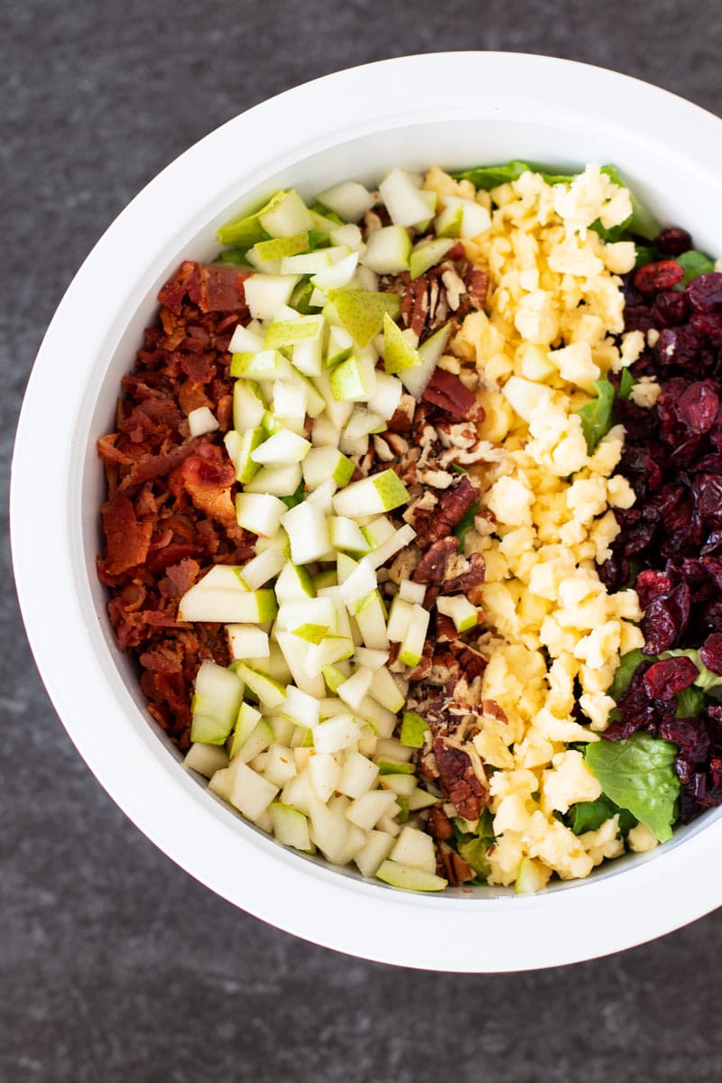 White bowl containing a salad with lettuce, bacon, pears, pecans, cheese, and dried cranberries.