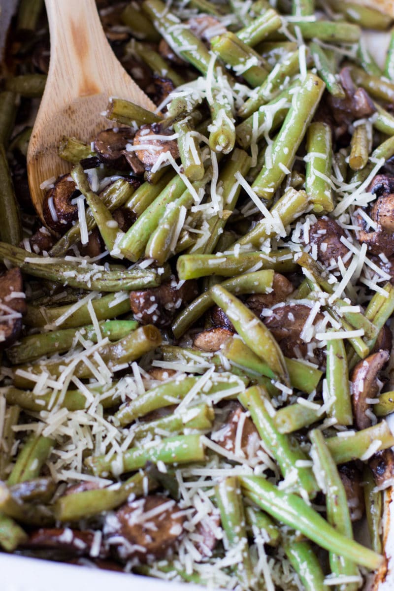 Baked green beans and mushrooms topped with Parmesan Cheese, wooden spoon in dish.