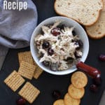 White bowl with a serving knife containing chicken salad topped with red grapes, crackers on table.