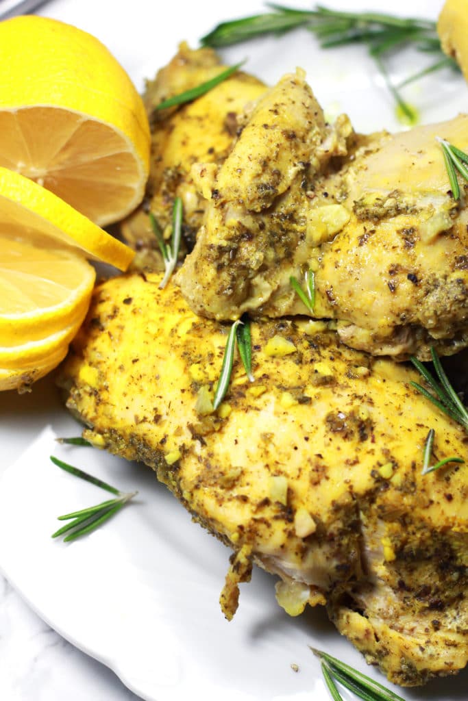 A white plate of three pieces of chicken covered in sprigs of herbs with lemon slices.