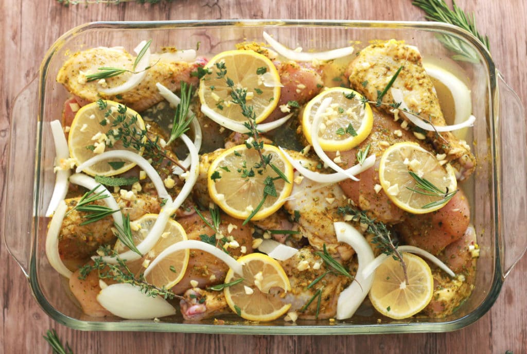 A dish of uncooked chicken covered with onions, sliced lemons, diced garlic, and sprigs of herbs.