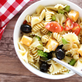White bowl containing pasta salad topped with tomatoes, peppers, cheese and broccoli, fork in bowl with red gingham napkin on brown table.