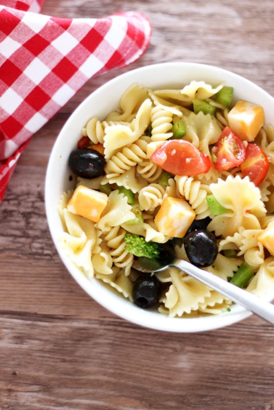 White bowl containing pasta salad topped with tomatoes, peppers, cheese and broccoli, fork in bowl with red gingham napkin on brown table.