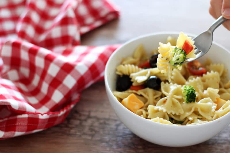 White bowl containing pasta salad topped with tomatoes, peppers, cheese and broccoli, fork with pasta, red gingham napkin on brown table.