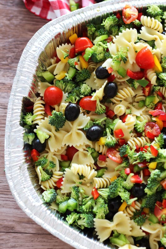 Silver pan containing pasta, broccoli, sweet peppers, tomatoes and black olives sitting on a brown table.