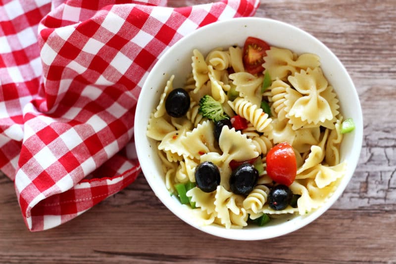 White bowl containing Italian Style Pasta Salad sitting on a brown table, red gingham napkin on table.