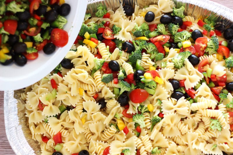 Silver pan containing Italian Style pasta salad with tomatoes, peppers, cheese and broccoli being poured in from a white bowl.