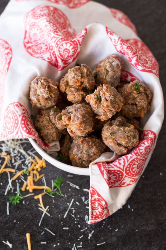 White bowl containing cooked sausage balls, parsley and cheese on counter.