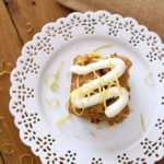 Lace white place containing a Carrot and Zucchini Bars with Lemon Cream Cheese Frosting sitting on a brown table, topped with lemon zest and fork on side of plate. Glass of milk in background.