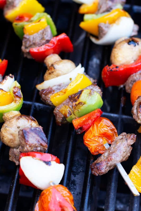 Steak and vegetables on three metal skewers on a grill. 