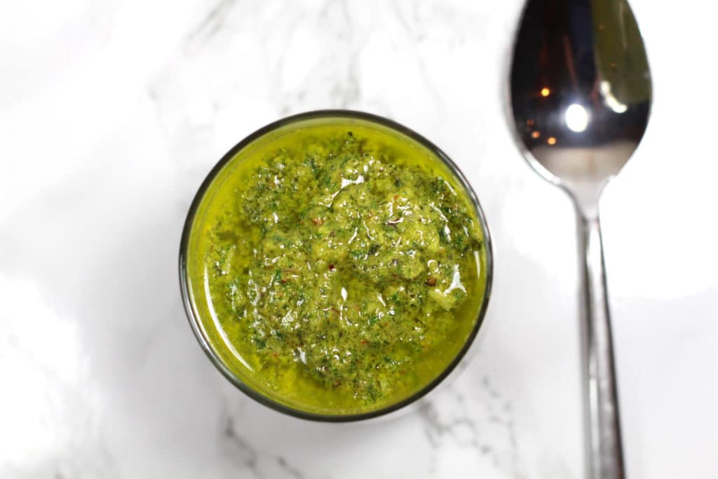 Jar of green dairy free pesto sauce on a white marble table, spoon on table.