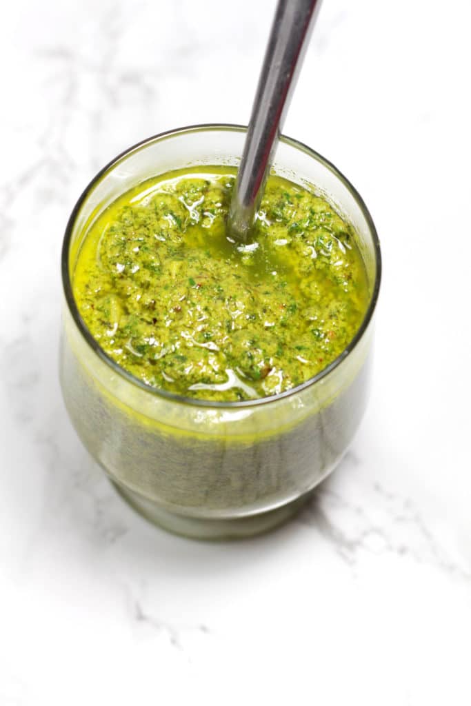 Jar of green dairy free pesto sauce on a white marble table, spoon in jar.