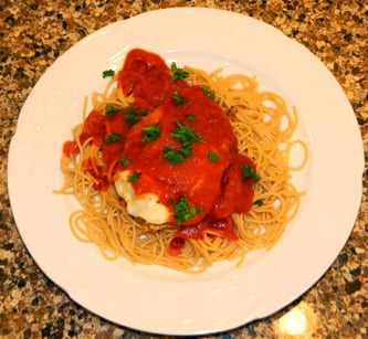 A white plate of spaghetti noodles topped with a chicken breast covered in marinara sauce.