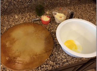 A pizza cooking stone, a white bowl with an egg, a red measuring cup of parmesan cheese, and riced cauliflower.