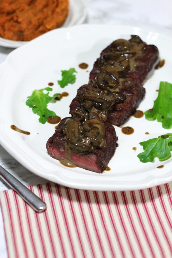 A white plate of steak and mushrooms drizzled with a creamy balsamic mushroom sauce.