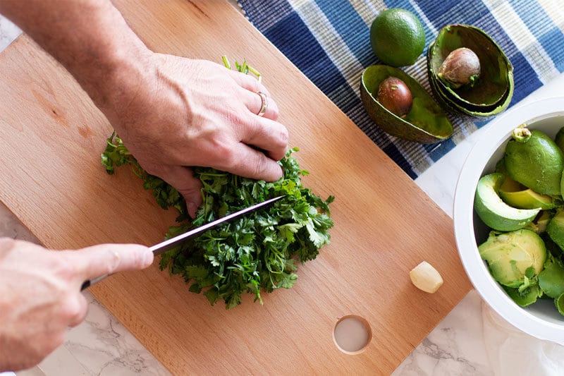 Man chopping cilantro with a knife, avocados and limes on table.