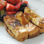 6 slices of french toast sitting on a white plate topped with maple syrup, side of fresh strawberries, grapes and blackberries.