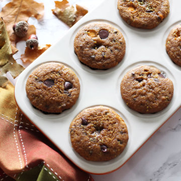 White muffin tin containing 6 Pumpkin Chocolate Chip muffins, napkin, leaves and acorns on table.