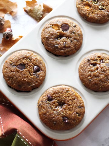 White muffin tin containing 6 Pumpkin Chocolate Chip muffins, napkin, leaves and acorns on table.