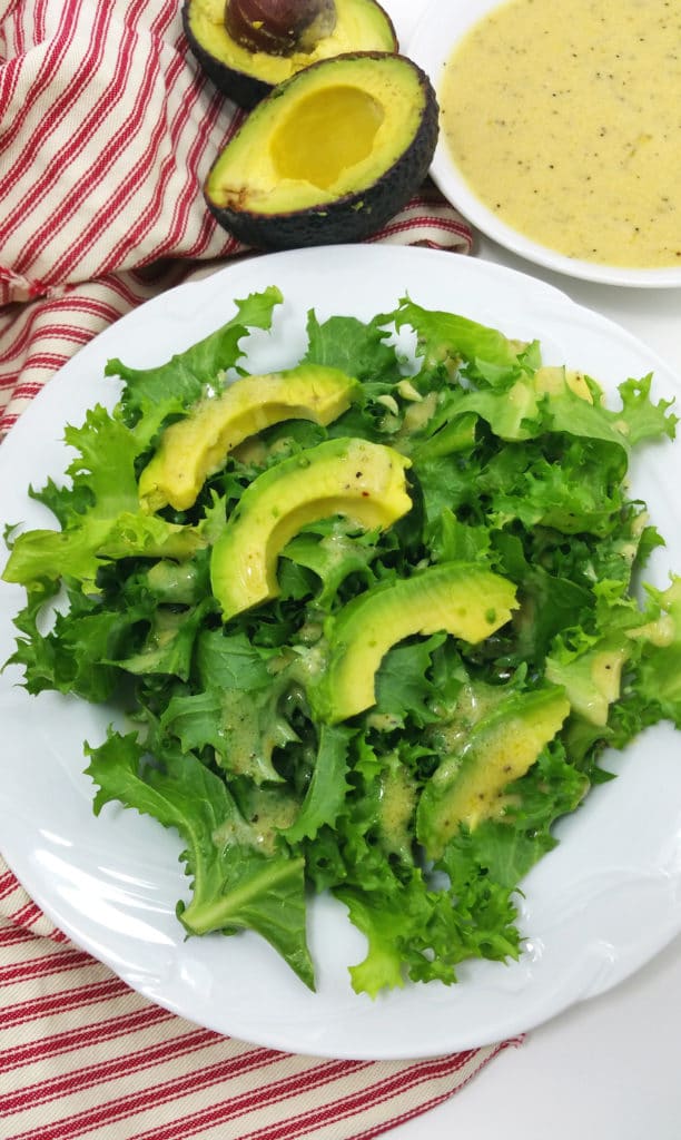 Endive Salad on a white plate, topped with fresh sliced avocado, dressing on side with red and white napkin on side. 