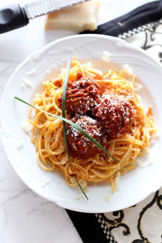 White plate containing spaghetti noodles topped with marinara sauce, 3 meatballs and parsley sitting on a white table, block of Parmesan cheese and grater.