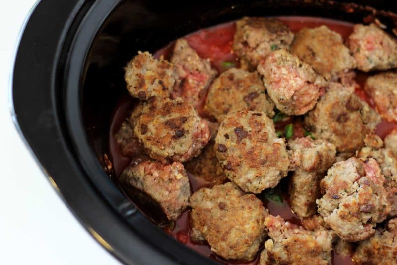 Black slow cooker containing seared meatballs sitting on top of red marinara sauce.