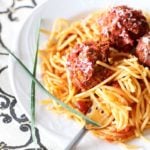 Three Meatballs sitting on top of spaghetti noodles with marinara sauce sitting on a white plate, fork and parsley.