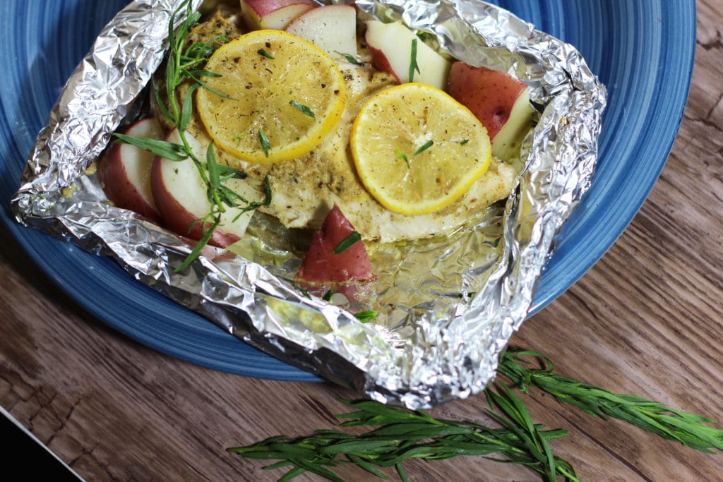 A close up of an aluminum foil tray filled with chopped potatoes, a chicken breast cover by two lemon slices on a table. 