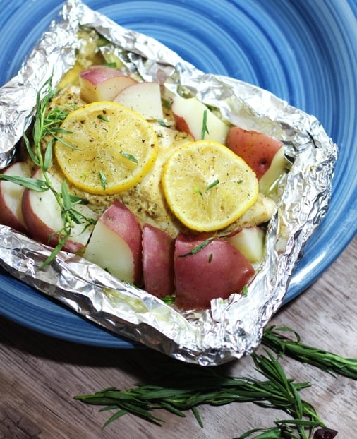 A close up of a blue plate with an aluminum foil tray filled with chopped potatoes, a chicken breast cover by two lemon slices. 
