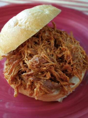 A red plate of pulled bar-b-que chicken on a kaiser roll.