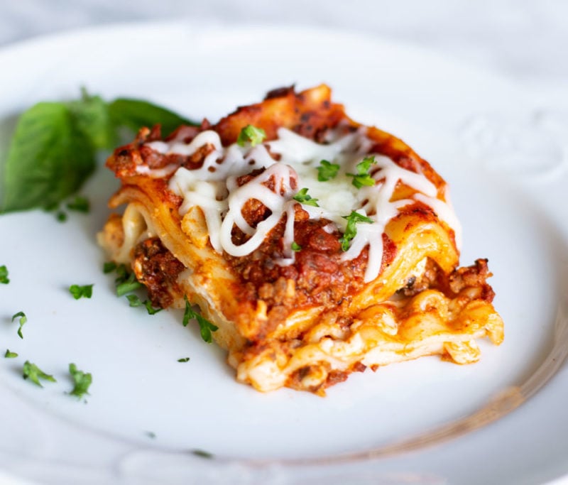 White plate containing a slice of cooked7 layered lasagna with an Italian meat sauce topped with melted mozzarella cheese and fresh basil.