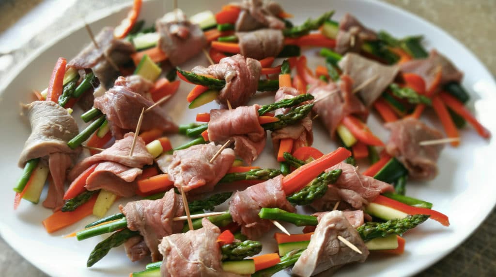 White serving dish containing 20 asian steak slices rolled up over asparagus, carrots, zucchini and secured with a toothpick.