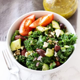 White bowl containing kale, cucumbers, feta cheese, Kalamata olive, and tomatoes, Greek salad dressing on table.