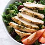 White bowl containing Greek Kale and Quinoa Salad, topped with a Greek Marinated chicken breast.