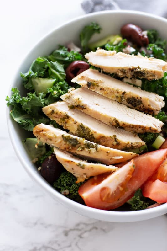 White bowl containing Greek Kale and Quinoa Salad, topped with a Greek Marinated chicken breast.