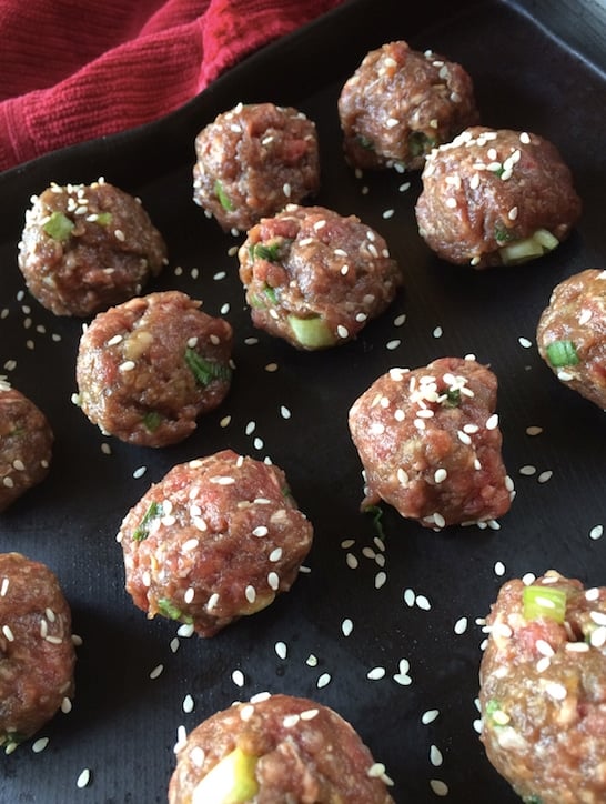 Rows of 12 uncooked ground beef meatballs stuffed with green onions, chopped garlic, and sprinkled with sesame seeds.