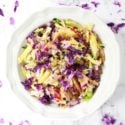 White bowl containing purple cabbage, squash, apple and celery, sitting on a white granite table, sprinkled with purple cabbage shavings.