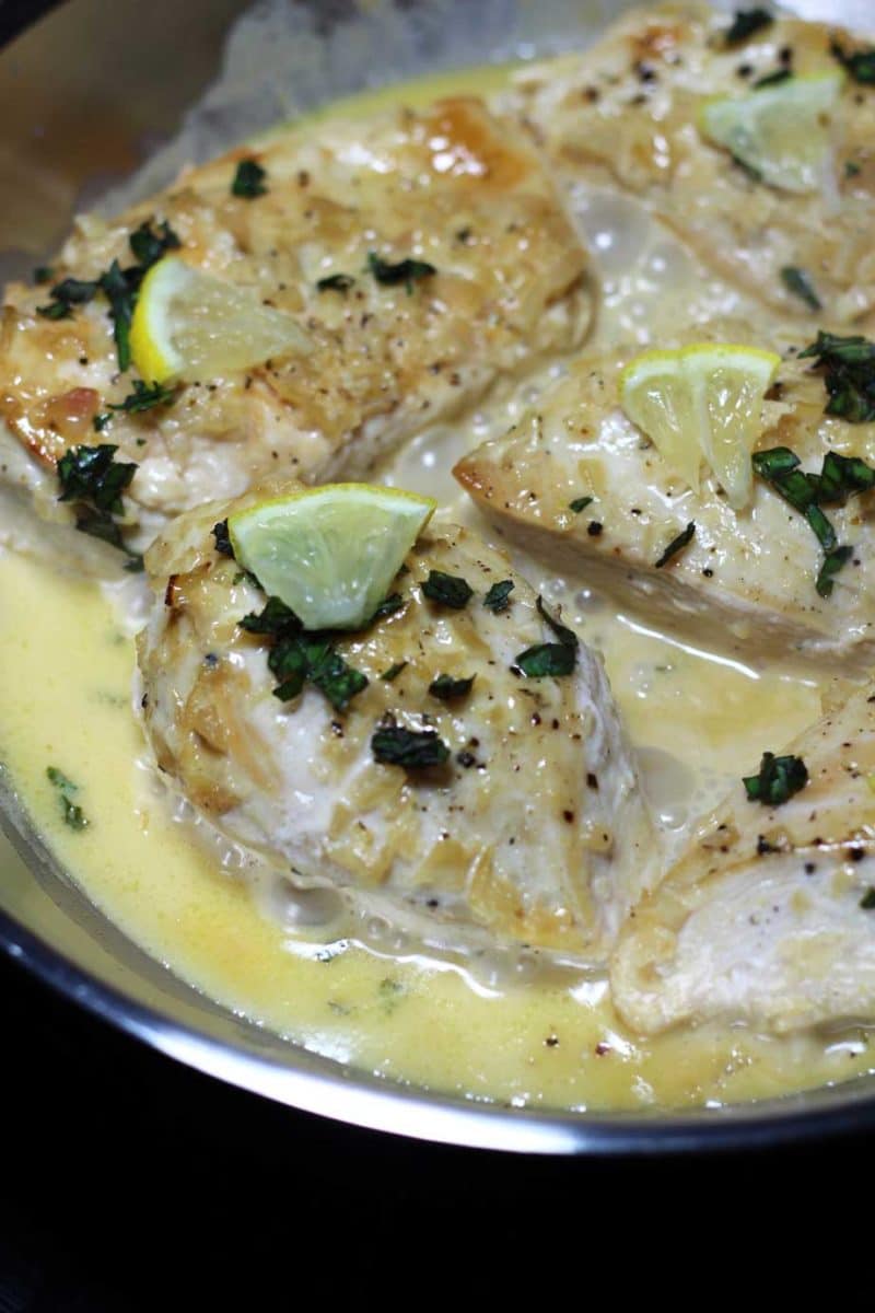 A close up of chicken smothered with a cream sauce and cut lemon on a plate. 