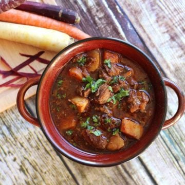 A bowl of beef stew with meat, carrots, and potatoes surrounded by carrots on a table.