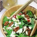 A wooden bowl of arugula salad topped with candied pecans, diced apples, and goat cheese crumbles.