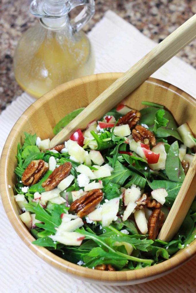 A wooden bowl of arugula salad topped with candied pecans, diced apples, and goat cheese crumbles.
